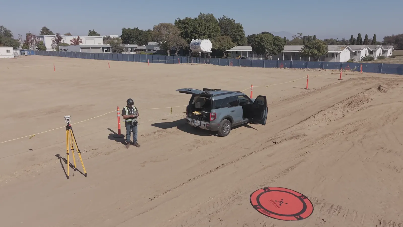 A crew member on a graded survey site with a GNSS rover on a tripod, a drone landing pad and a field vehicle.