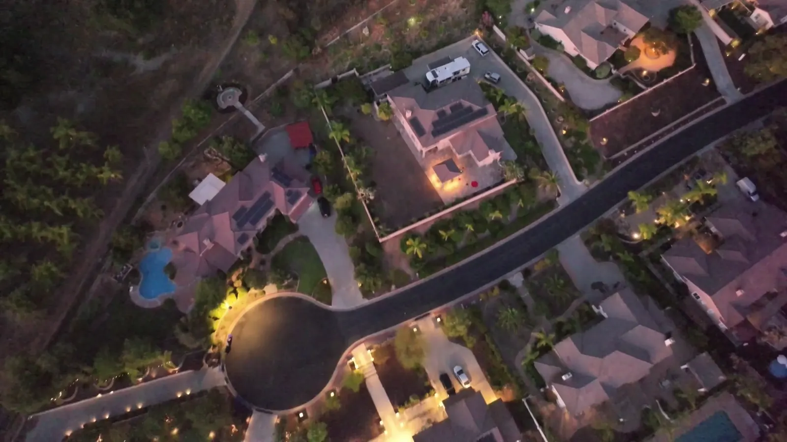A top-down drone view of a residential cul-de-sac at dusk, with rooftops, pools and lit driveways.