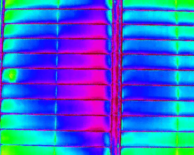 A thermal drone image of a rooftop solar array: rows of panels in blue and green, with one warm cell showing yellow at the left edge.