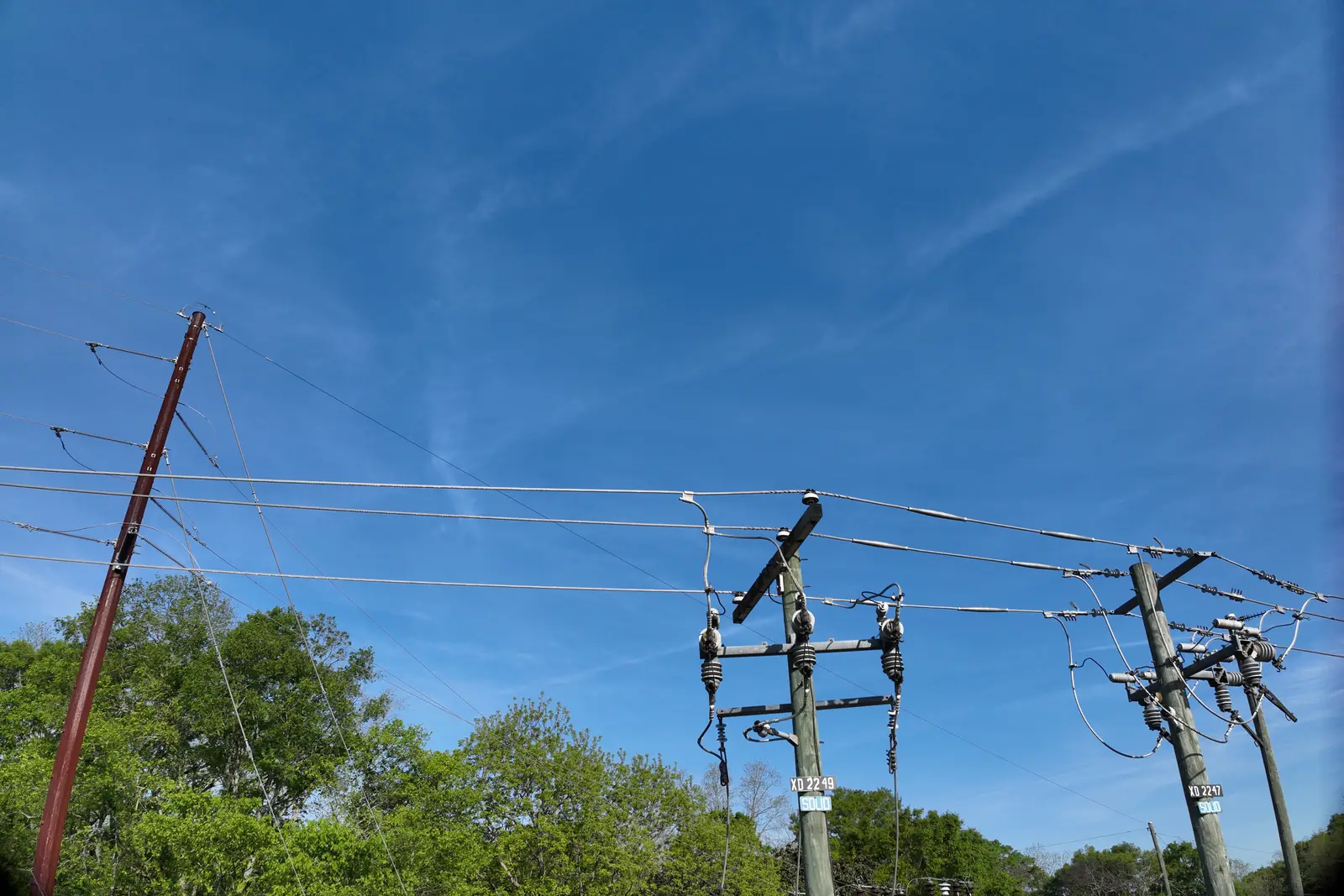 A distribution line from the air: poles, crossarms and conductors against a blue sky above the tree line.