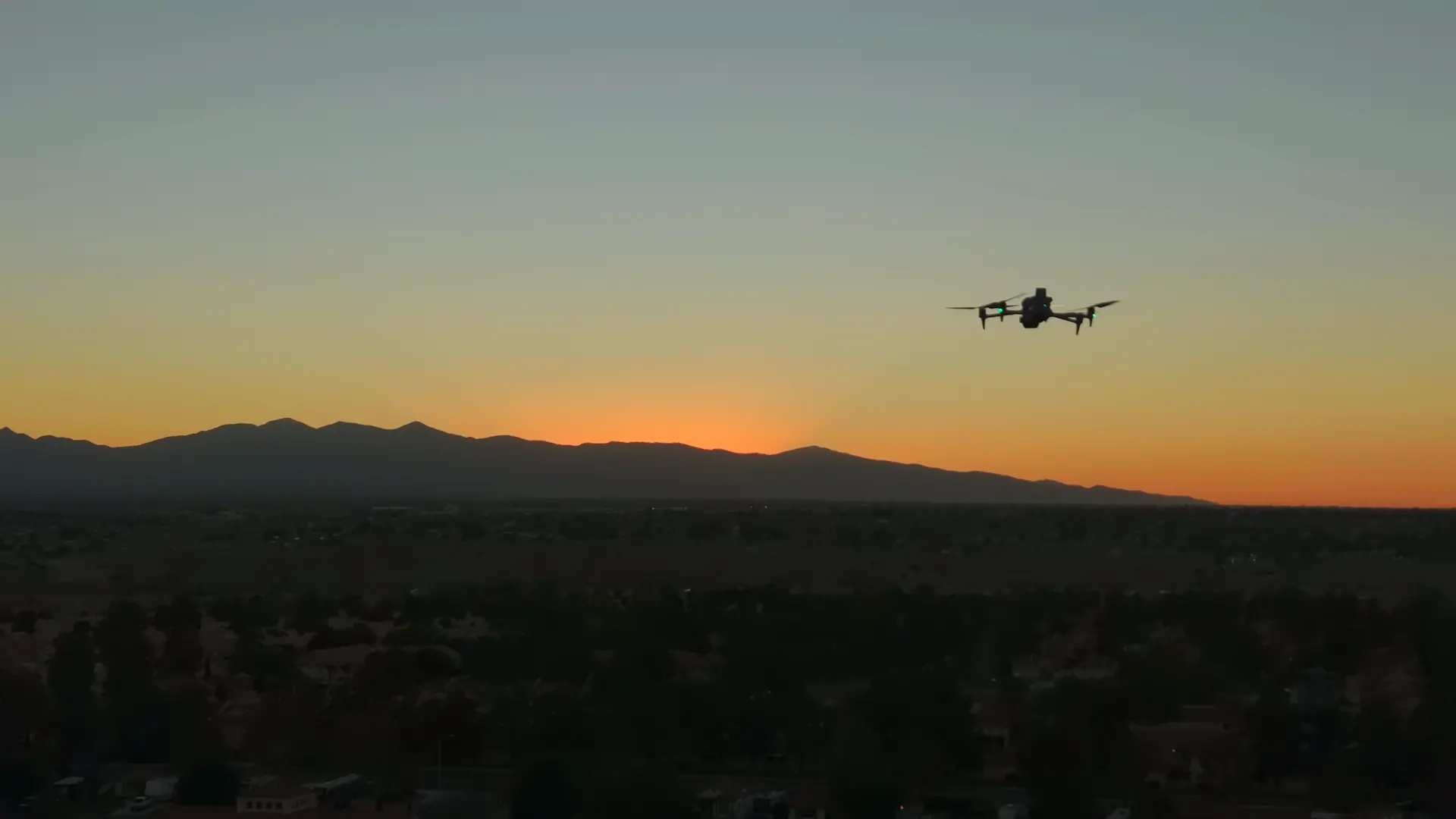A drone hovering at sunset in front of a mountain ridge, with rooftops and trees far below.